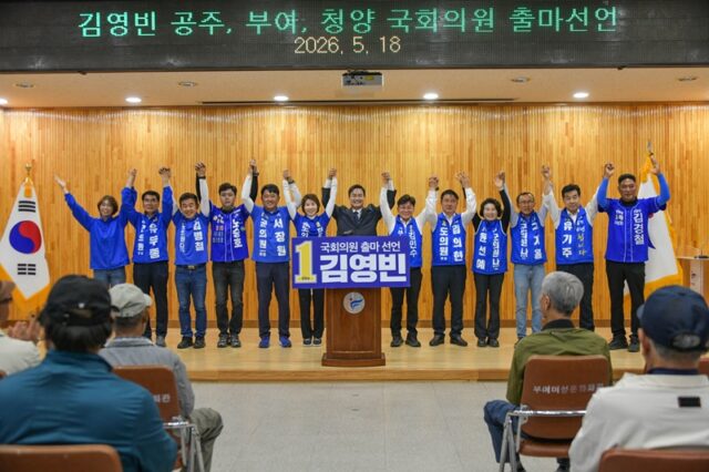 2026년 5월 18일, 부여군여성문화회관에서 열린 국회의원 보궐선거 출마 기자회견에서 더불어민주당 김영빈 후보와 부여 지역 시·도의원 출마자들이 손을 맞잡고 번쩍 들어 올리며 참석자들에게 인사하며 원팀임을 강조하고 있다.